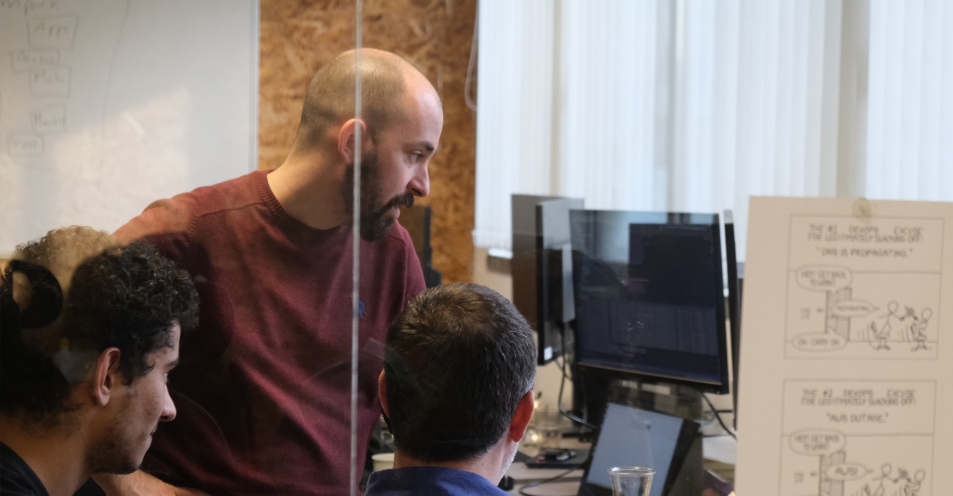 Three young people look focused at a computer screen while solving a programming problem in an office full of computers.